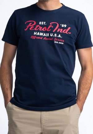 Uomo che indossa una t-shirt blu navy con testo rosso e bianco che recita Petrol Ind. Hawaii U.S.A. Offroad Rental Service, abbinata a pantaloni beige.