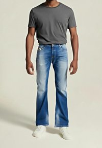 Homme portant un jean bootcut bleu délavé, un t-shirt gris à manches courtes et des baskets blanches, debout devant un fond uni.