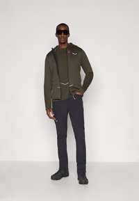 Ελιά πρασινής ζακέτα με φερμουάρ πάνω από σκούρο πουκάμισο, σε συνδυασμό με σκούρους παντελόνι και μαύρες μπότες. Διαθέτει ανακλαστικές λεπτομέρειες και κουκούλα.