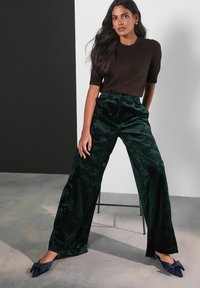 Dark green velvet wide-leg pants with a textured floral pattern, paired with a brown fitted top and navy pointed flats with bow accents.