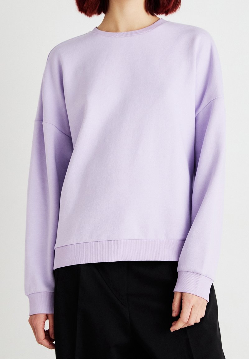 Personne portant un sweat-shirt violet clair à manches longues et col rond, ainsi qu'un pantalon noir, debout devant un fond blanc.
