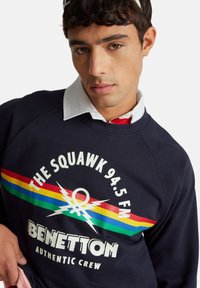 Mladý muž s krátkými tmavými vlasy, oblečený v námořnicky modré mikině s barevnými pruhy a nápisem "THE SQUAWK 94.5 FM BENETTON AUTHENTIC CREW".