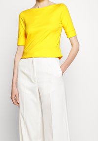 Gul kortärmad topp med båtringning och figurnära design, kombinerad med vita vida byxor. Slät, elastisk tyg.