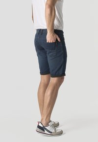 Férfi sötétkék felhajtott farmer rövidnadrágban, fehér pólóban és fehér tornacipőben, egyik kezével a hátsó zsebében áll.