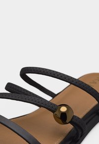 Gros plan d'une sandale noire à brides fines tressées avec une perle métallique ronde brillante sur une semelle couleur cuir.