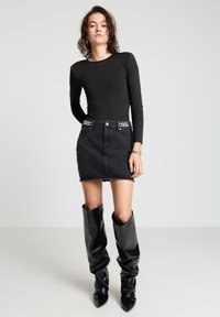 Schwarzer Langarm-Body kombiniert mit einem schwarzen Denim-Minirock mit ausgefranstem Saum und silbernen Nietenakzenten am Bund. Schwarze kniehohe Stiefel.