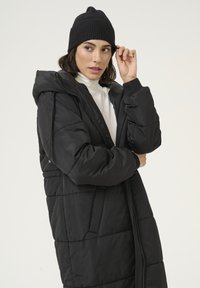 Black padded long coat with a hood, featuring a zip closure and side pockets. Worn with a white turtleneck and a black knit beanie.