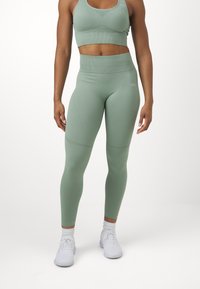 Leggings de sport vert clair avec une taille haute, fabriqués en tissu extensible. Comprend une texture lisse et un design ajusté. Associés à des chaussures blanches.