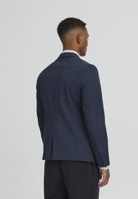 Tengerészkék zakó, amely testre szabott fazonnal, kiszögelős hajtókákkal és egyetlen szellőzőnyílással rendelkezik a hátán; sima anyag és gombos mandzsetta.