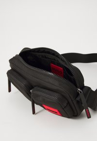 Schwarze Stoff-Schultertasche mit zwei Reißverschlussfächern, einem roten Logo-Patch und verstellbarem Tragegurt. Glatte Oberfläche mit dezentem Glanz.