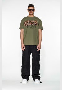 Olivengrønn t-skjorte med "Mugonzales Studios" grafikk, kombinert med svarte cargobukser. Hvite sneakers og mørke solbriller fullfører stilen.