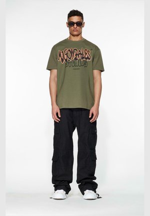 Olivengrønn t-skjorte med "Mugonzales Studios" grafikk, kombinert med svarte cargobukser. Hvite sneakers og mørke solbriller fullfører stilen.