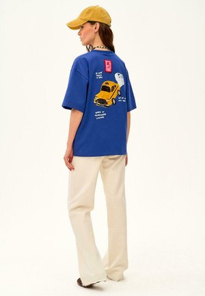 Kobieta w niebieskiej, luźnej koszulce z żółtą grafiką taksówki i napisem na plecach, beżowe spodnie, żółta czapka i naszyjnik, zwrócona w lewo.