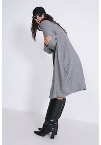 Grijze, gestructureerde oversized cardigan met lange mouwen, gecombineerd met zwarte, kniehoog, met hakken laarzen en zwarte zonnebril, afgewerkt met een zwarte pet.