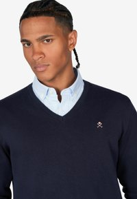 Maglione a V blu navy con una texture liscia, indossato sopra una camicia colletto azzurro chiaro. Presenta un piccolo logo di rosa e croce sul petto.