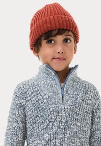 Un maglione lavorato a maglia blu e bianco con colletto alto e mezza zip, abbinato a un berretto a coste in tonalità ruggine.