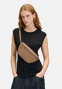 Tan-farbenes Leder-Crossbody-Bag mit glatter Textur, Reißverschluss und verstellbarem Tragegurt, getragen über einem schwarzen gerippten Oberteil.