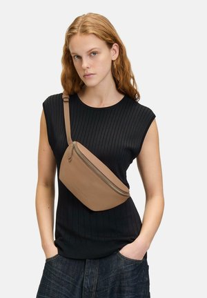 Tan-farbenes Leder-Crossbody-Bag mit glatter Textur, Reißverschluss und verstellbarem Tragegurt, getragen über einem schwarzen gerippten Oberteil.