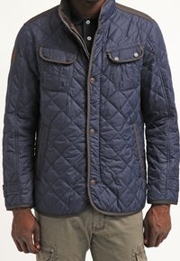 Marineblå quiltet jakke med knaplukning, to brystlommer og sidelommer; har kontrasterende brun kant langs kanterne.