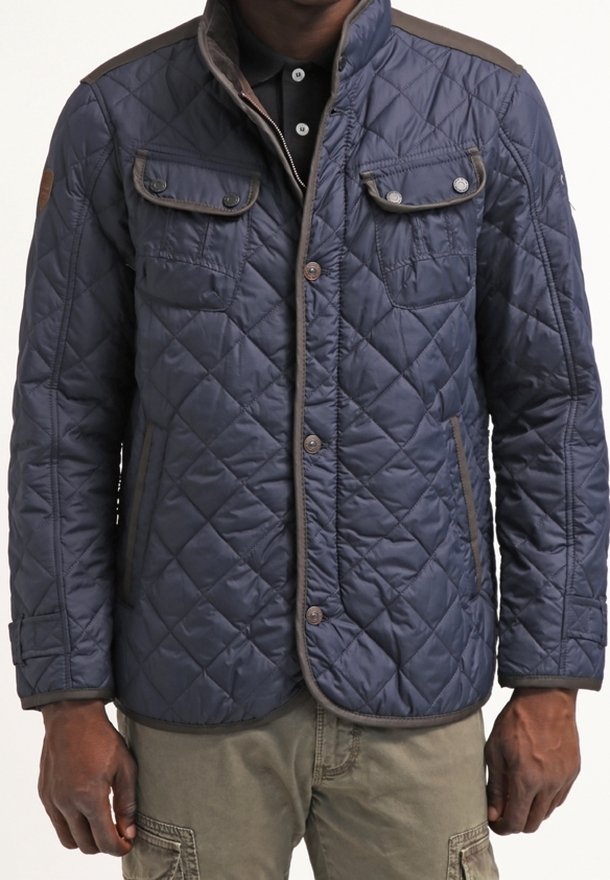 Marineblå quiltet jakke med knaplukning, to brystlommer og sidelommer; har kontrasterende brun kant langs kanterne.