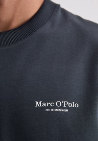Granatowa bluza wykonana z teksturowanej tkaniny, z prążkowanym, okrągłym dekoltem oraz białym logo z napisem "Marc O'Polo" i "EST. IN STOCKHOLM."