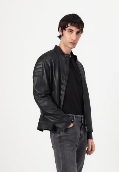 Veste en cuir noir avec des accents côtelés sur les épaules, fermeture à glissière à l'avant et col montant. Portée sur une chemise noire et un jean gris.