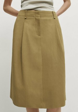 Femme portant une jupe beige longueur genou avec passants de ceinture et un haut blanc sans manches, mains posées sur les hanches devant un fond uni.