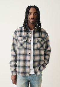 Camicia di flanella a quadri nei toni del nero e grigio, con chiusura frontale a bottoni, due tasche sul petto e vestibilità casual su una t-shirt bianca.