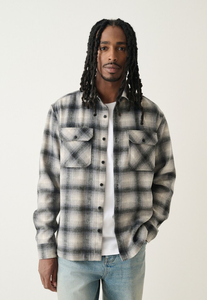 Camicia di flanella a quadri nei toni del nero e grigio, con chiusura frontale a bottoni, due tasche sul petto e vestibilità casual su una t-shirt bianca.