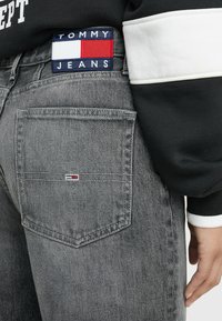 Los jeans de mezclilla gris presentan un bolsillo trasero con un pequeño parche de logo de Tommy Jeans en rojo, blanco y azul. Los jeans se combinan con una camiseta negra.