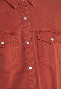 Camicia di tessuto color ruggine con chiusura a bottoni, colletto a punta e due tasche sul petto con bottoni bianchi. Texture liscia.