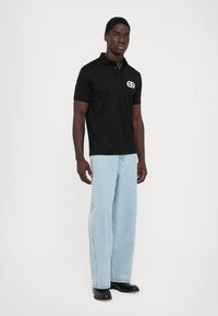 Tricou polo negru cu mânecă scurtă și guler, având un logo alb pe piept. Asortat cu blugi largi de culoare albastră deschisă și pantofi negri.