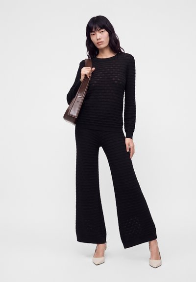 Femme portant un haut noir texturé à manches longues et un pantalon large, tenant un sac à épaule marron, debout avec une expression neutre.