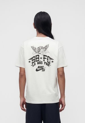 Osoba z długimi ciemnymi włosami ubrana w białą koszulkę Nike SB z grafiką anioła i tekstem "SB FC La Mas Fina" na plecach.