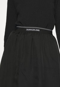 Musta hame, jossa on ryppyinen pinta, leveällä vyötärönauhalla, jossa on valkoiset tekstit "CALVIN KLEIN JEANS", korostettuna ohuilla valkoisilla raidoilla.