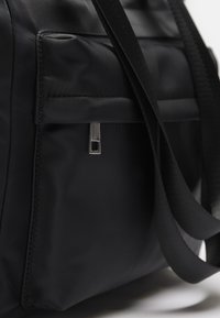 Black nylon bag featuring a smooth texture, zippered pocket with a rectangular pull, and flat, strong carrying straps.