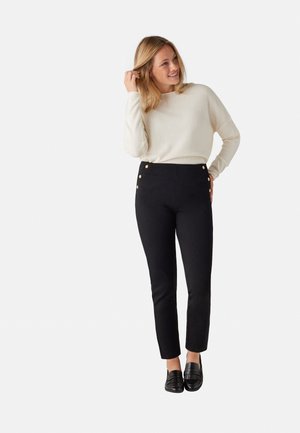 Pantalones negros ajustados con cuatro botones decorativos dorados en el lado, combinados con un suéter de punto crema y mocasines negros.