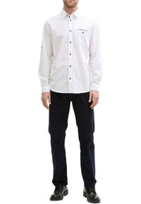 Chemise blanche à manches longues avec col boutonné, poche poitrine et boutons marron, associée à un pantalon noir et des chaussures noires.