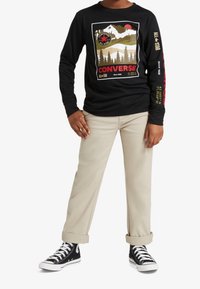 Schwarzes langärmeliges T-Shirt mit Berggrafik und dem Schriftzug "CONVERSE". Kombiniert mit hellbeigen Hosen und schwarzen High-Top-Sneakern.