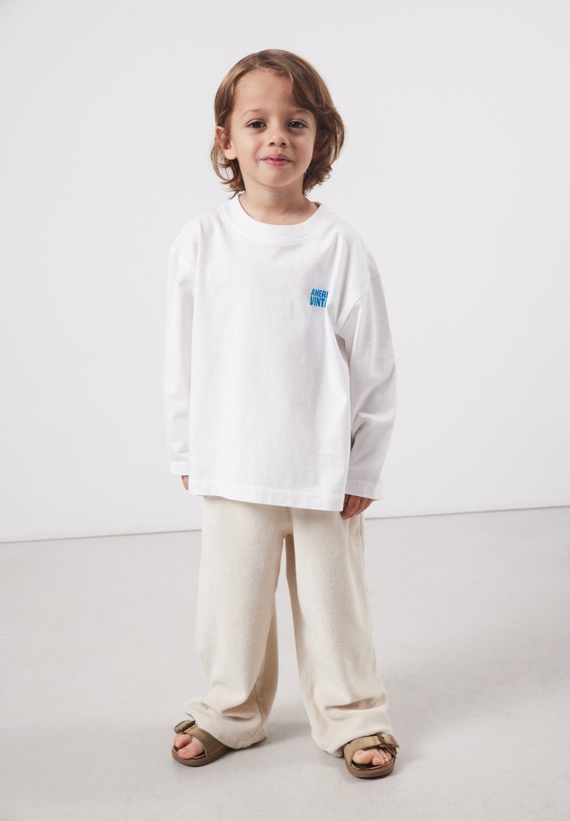 Jeune enfant aux cheveux mi-longs portant une chemise blanche à manches longues trop grande, un pantalon beige et des sandales marron sur un fond uni.