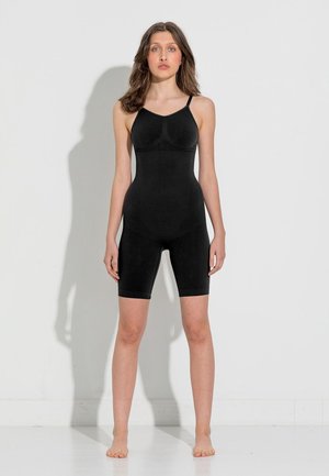 Vrouw staat blootsvoets tegen een witte muur en draagt een zwart, mouwloos corrigerend kledingstuk met korte pijpen.