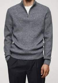 Pull gris en maille côtelée avec col zippé, manches longues et coupe décontractée, associé à un pantalon foncé. Texture douce.