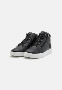 Zapatillas altas sintéticas negras con cordones en la parte frontal, cremallera lateral y suela de goma blanca. Textura suave con detalles de diseño mínimos.