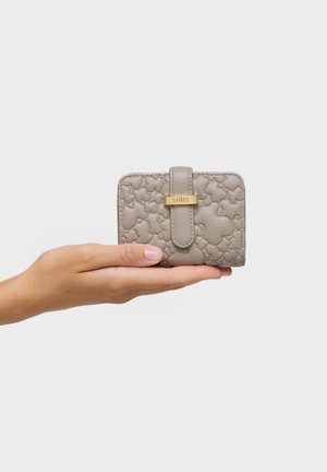 Mano sosteniendo una pequeña billetera beige con patrones de osos en relieve y un broche dorado grabado con "TOUS" sobre un fondo liso.