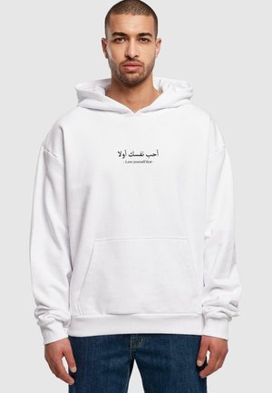 White hoodie made of soft fabric, featuring a kangaroo pocket. Text in Arabic and English reads "Love yourself first." Casual fit with ribbed cuffs.