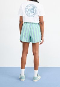 Weißes T-Shirt mit runder blauer Grafik auf dem Rücken, kombiniert mit leichten grün-blauen gestreiften Shorts. Getragen mit weißen Socken und türkisfarbenen Sneakers.