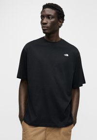 Νέος άνδρας με κοντά σγουρά μαλλιά που φοράει μαύρο t-shirt The North Face και καφέ παντελόνι, κοιτάζοντας ελαφρώς στην πλευρά με τα χέρια στις τσέπες.