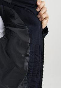 Interior de chaqueta azul marino, con un forro negro suave y cierre con cremallera. Costuras visibles y detalle de bolsillo en el lado izquierdo.