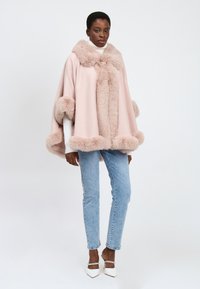 Kvinde iført en lys rosa kappe med pelskant over en hvid turtleneck, lyseblå jeans og hvide spidse hæle, stående foran en ensfarvet baggrund.