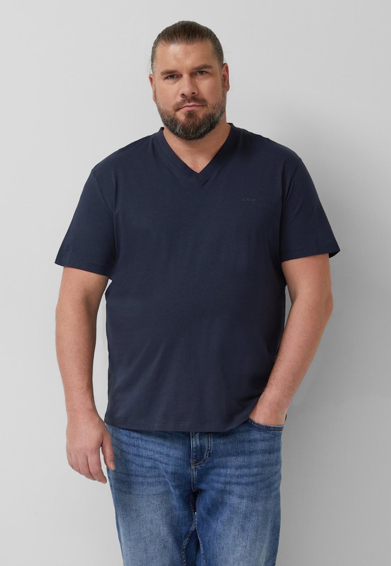Homme barbu aux cheveux attachés, portant un t-shirt bleu marine à col en V et un jean bleu, debout avec une main dans la poche devant un fond uni.